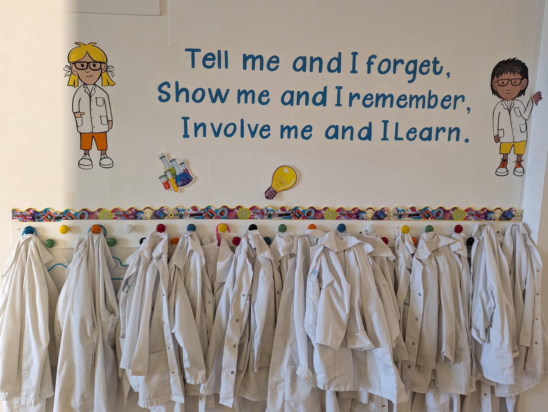 Mini Professors lab coats hanging beneath the quote: tell me and I forget, show me and I remember, involve me and I learn